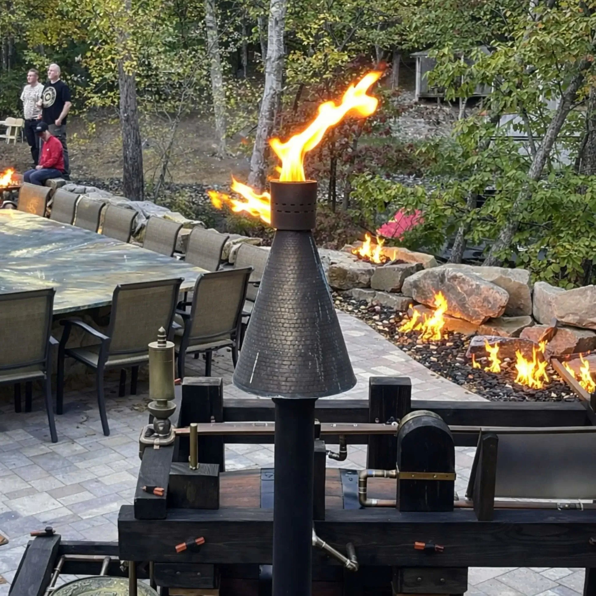 HPC TK Torch Copper Head and Post Lifestyle 7 installed near a large outdoor dining table and linear fire features, with multiple flames integrated into a stone landscape wall, highlighting how the torch complements luxury outdoor kitchens and entertainment areas.