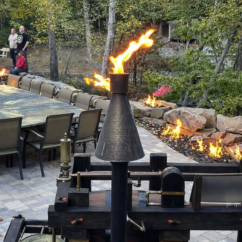 HPC TK Torch Copper Head and Post Lifestyle 7 installed near a large outdoor dining table and linear fire features, with multiple flames integrated into a stone landscape wall, highlighting how the torch complements luxury outdoor kitchens and entertainment areas.