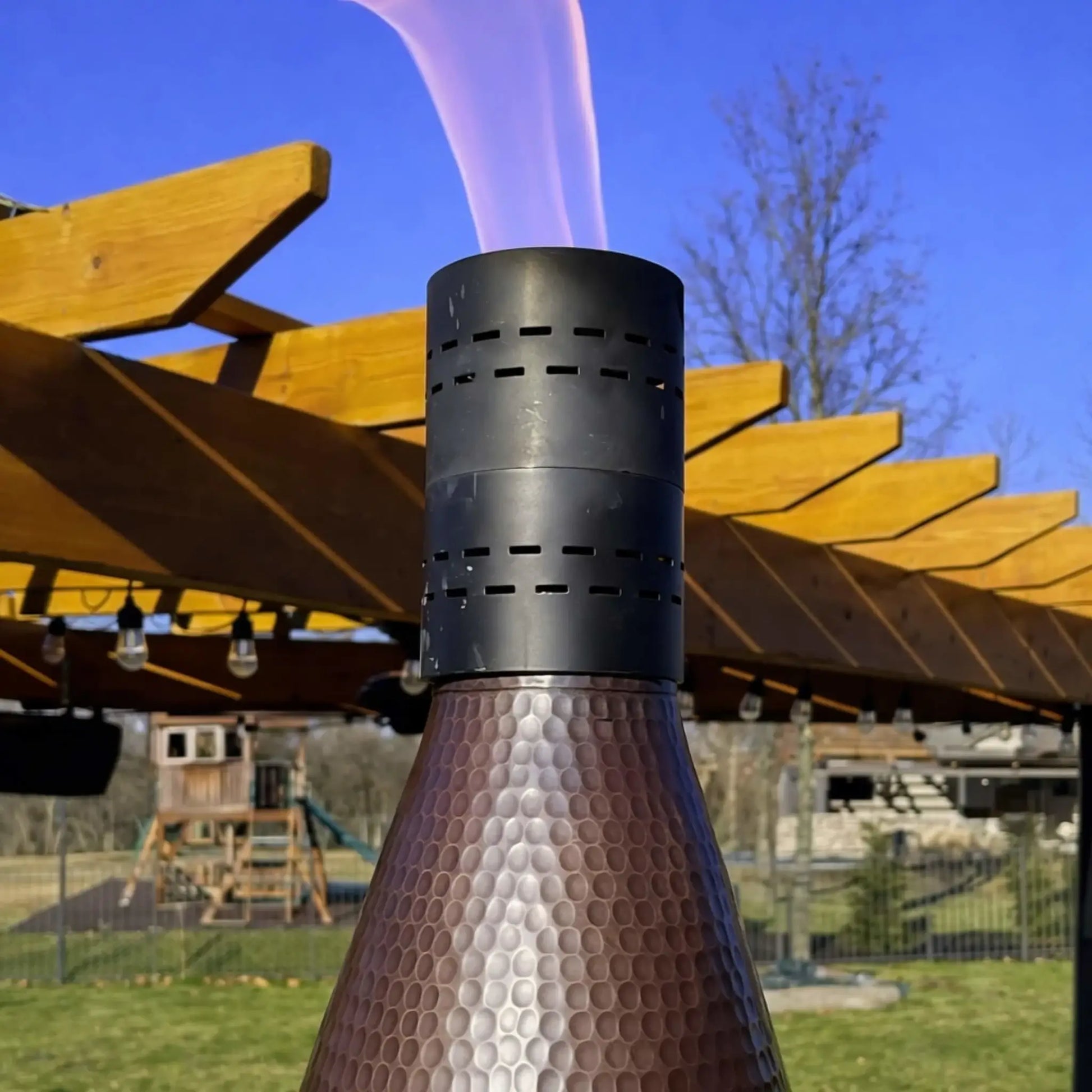 HPC TK Torch Copper Head and Post Lifestyle 8 close-up showing the hammered copper torch head beneath a pergola with string lights, with flame rising from the vented top, emphasizing the detailed texture and premium finish of the outdoor gas torch.