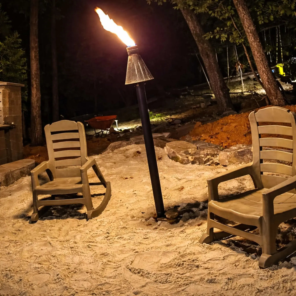HPC TK Torch Copper Head and Post Lifestyle 9 featuring a lit torch placed between two outdoor chairs on a sandy seating area at night, surrounded by trees and soft landscape lighting, creating a cozy fire-lit gathering space.