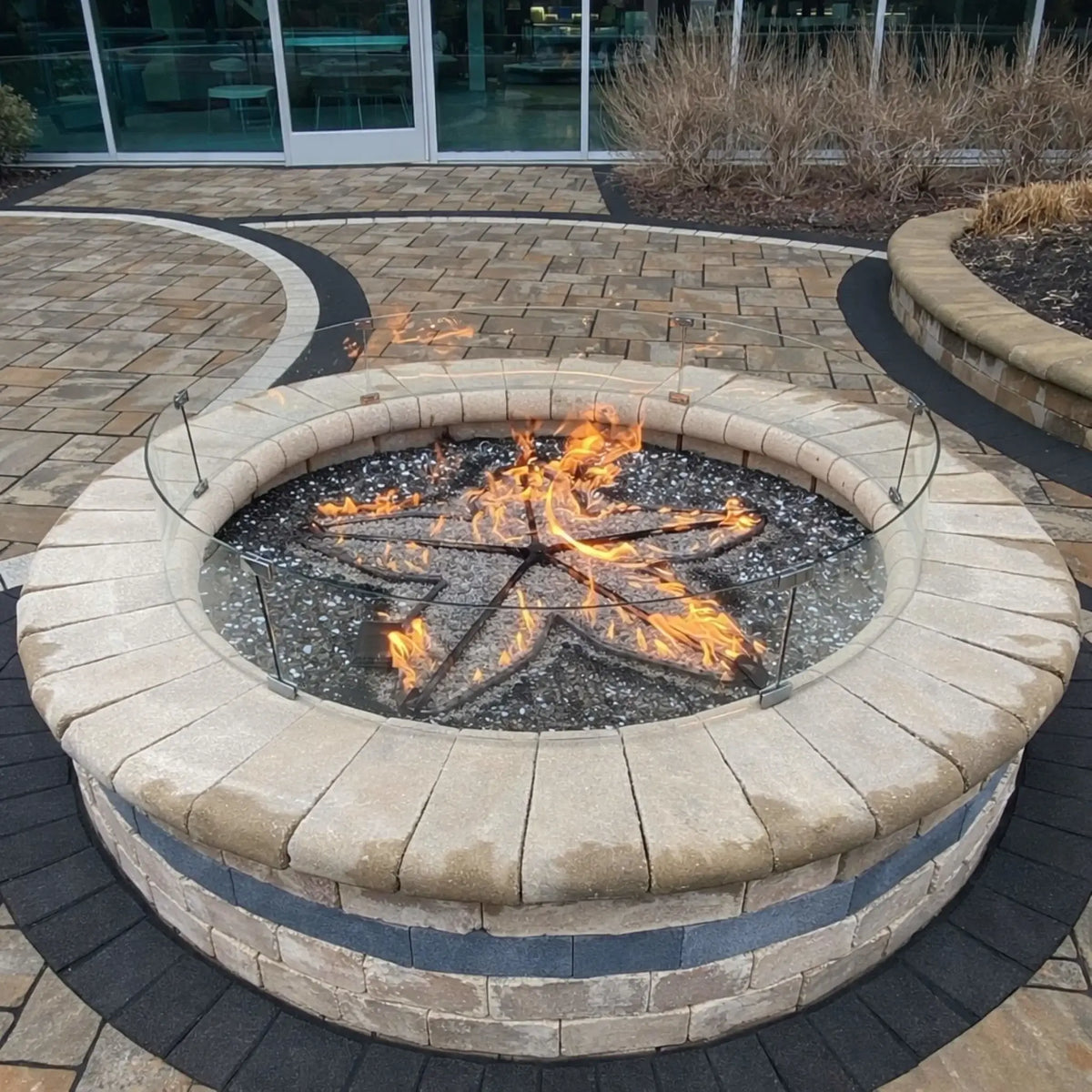 Close-up of a round outdoor gas fire pit with a stainless steel star burner and black fire glass, surrounded by a clear tempered glass wind guard. The fire pit is built with stacked stone and a smooth cap, set within a modern paver patio featuring decorative curved inlays near a glass building entrance.
