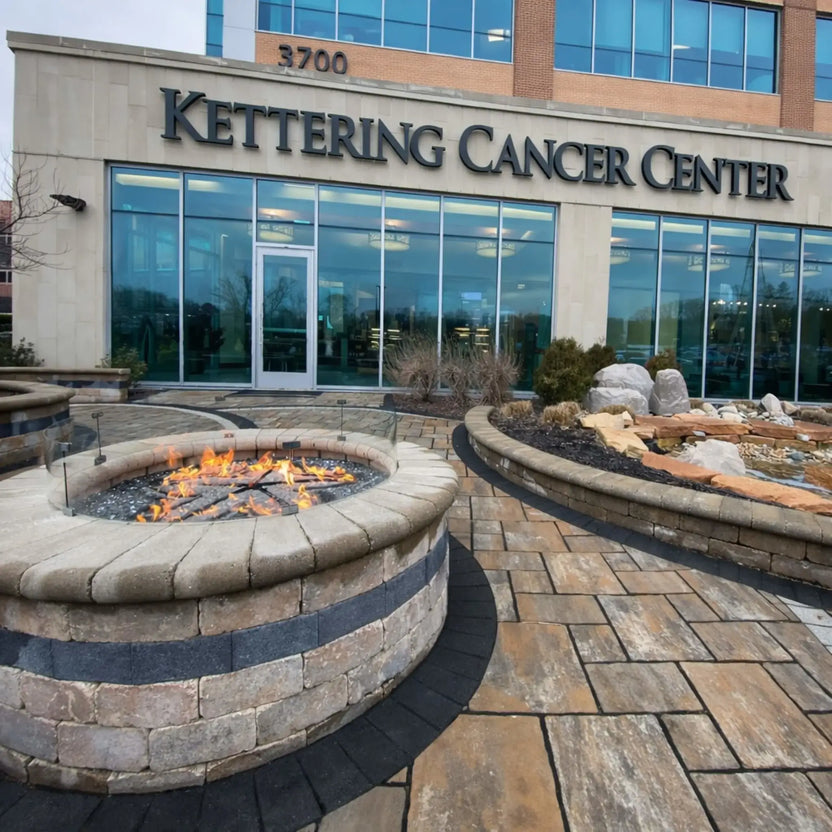 Wide view of a commercial outdoor plaza featuring a circular stone fire pit with a tempered glass wind guard and active flame. The fire feature sits in front of a modern building with large windows and signage reading “Kettering Cancer Center,” surrounded by decorative landscaping and patterned pavers.