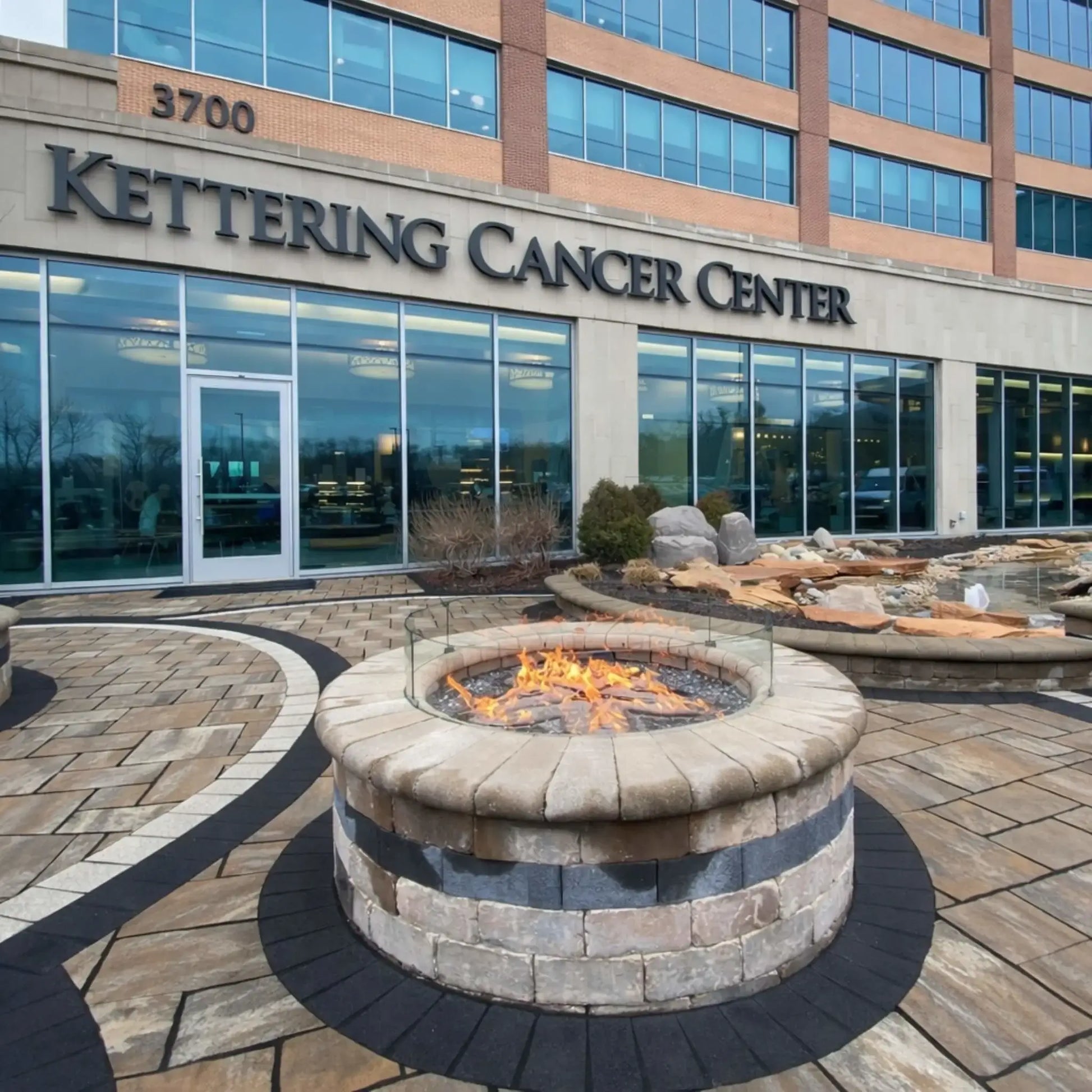 Angled perspective of a round gas fire pit with glass wind guard installed in a spacious paved courtyard outside Kettering Cancer Center. The fire pit features a stone veneer base and black accent band, complemented by nearby rock landscaping and curved seating walls.
