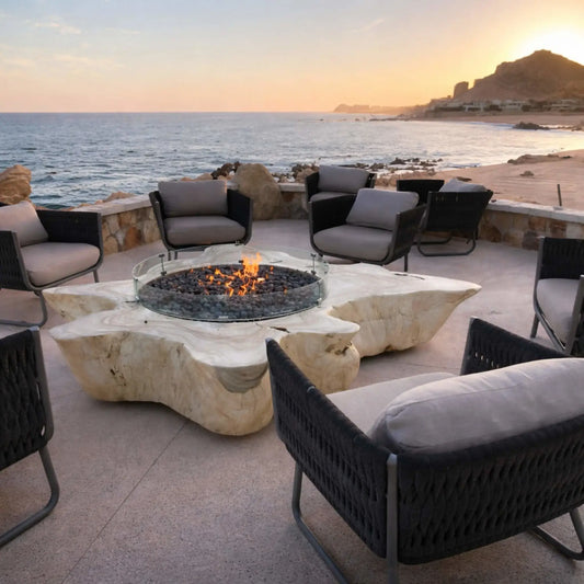 Luxury oceanfront patio featuring a natural stone fire table with a round tempered glass wind guard protecting a central gas flame. Surrounded by modern cushioned lounge chairs, the setting overlooks a rocky coastline and calm ocean at sunset, creating a warm, inviting outdoor gathering space.