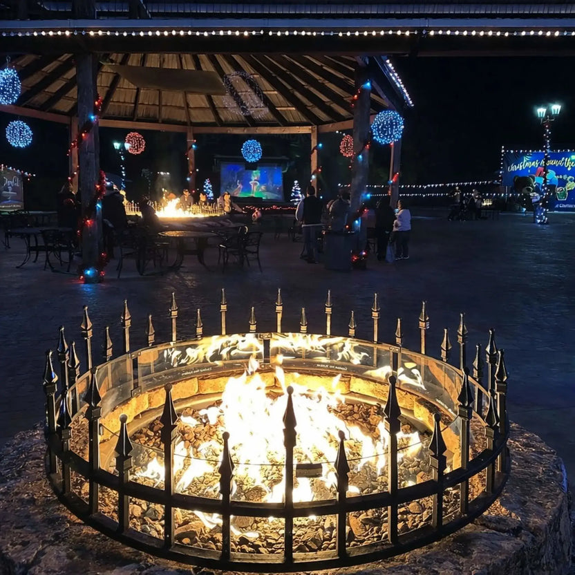 Nighttime outdoor pavilion with a circular fire pit enclosed by a decorative metal guard and glass wind barrier. The fire glows brightly as people gather nearby under string lights and festive decorations, creating a cozy, social atmosphere in a public plaza setting.