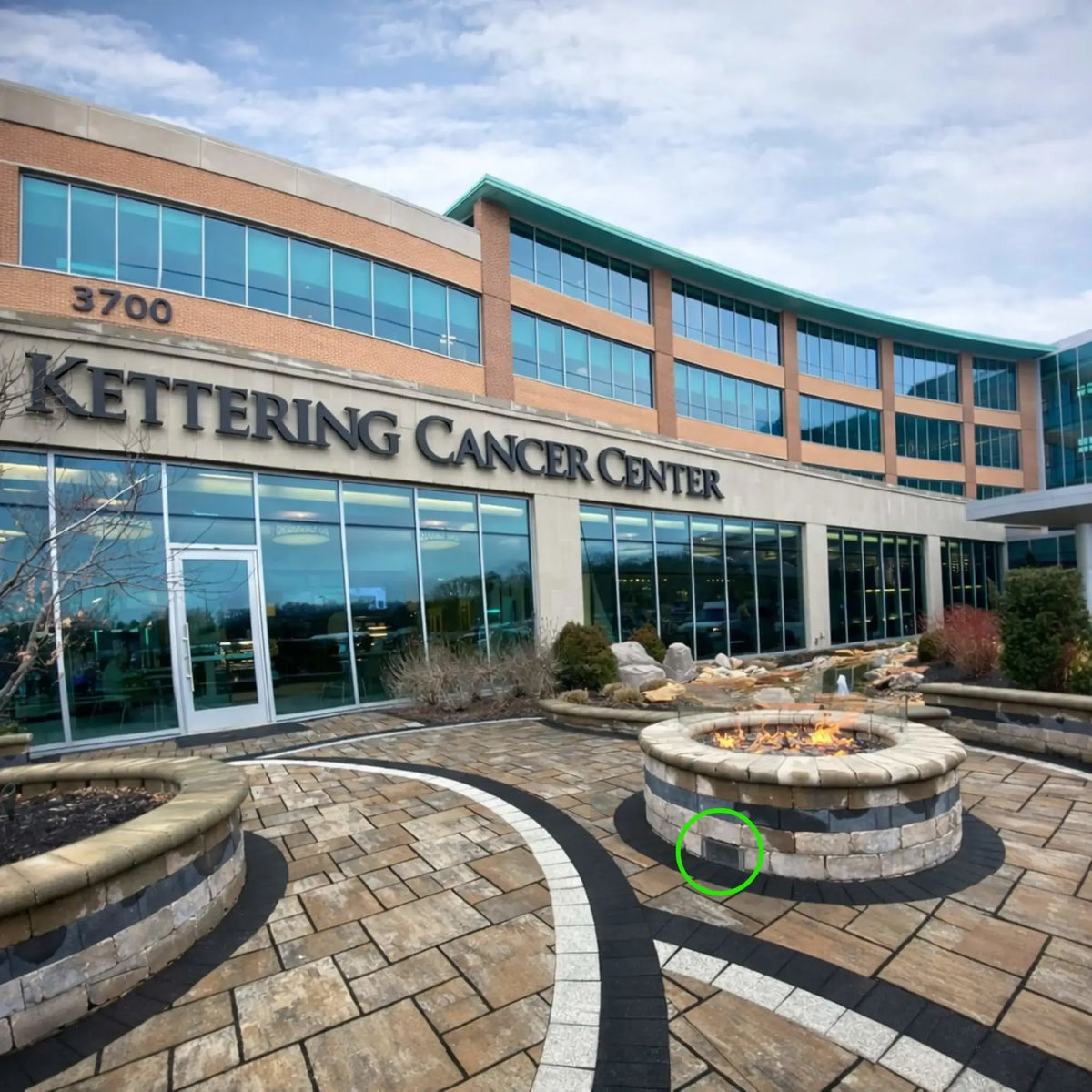 Outdoor courtyard at a medical campus featuring a large stone gas fire pit with flame glass burning in the center. A stainless steel ventilation grille is installed on the base of the fire pit enclosure to allow airflow and gas ventilation, helping ensure safe operation of the fire pit in commercial outdoor installations.