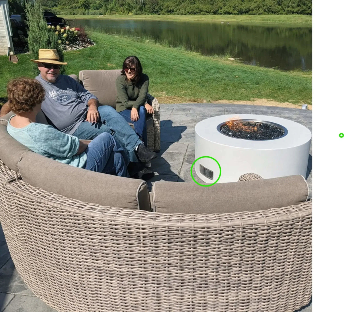 Friends relaxing around a round outdoor gas fire pit with black fire glass on a lakeside patio. The fire pit enclosure includes a stainless steel ventilation grille that allows air circulation and gas ventilation inside the structure, helping maintain safe operation of the gas fire feature.