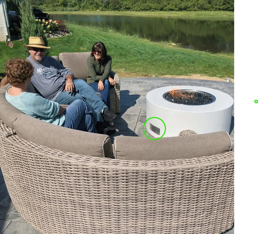 Friends relaxing around a round outdoor gas fire pit with black fire glass on a lakeside patio. The fire pit enclosure includes a stainless steel ventilation grille that allows air circulation and gas ventilation inside the structure, helping maintain safe operation of the gas fire feature.