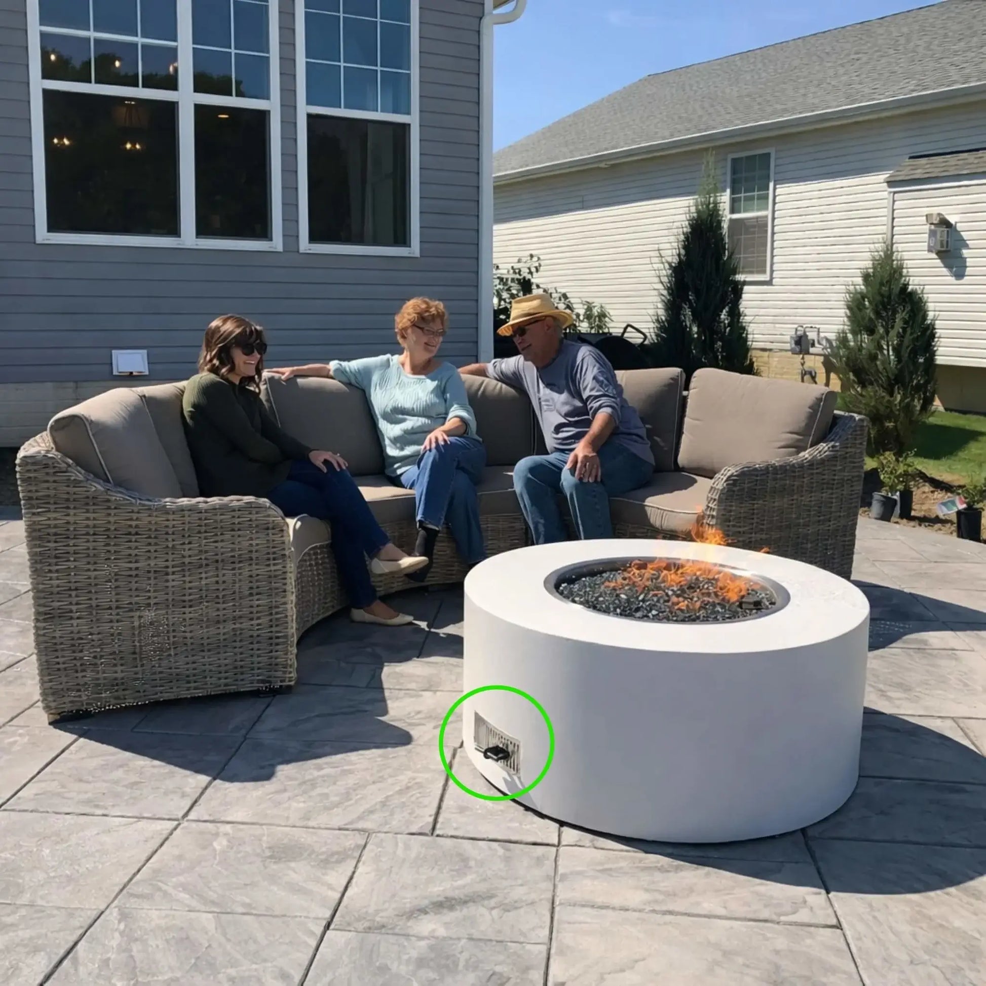 Outdoor patio seating area with wicker sofa and chairs arranged around a modern round concrete gas fire pit. A stainless steel ventilation grille is visible on the fire pit enclosure to allow airflow and ventilation for the gas burner system while guests gather comfortably around the fire.