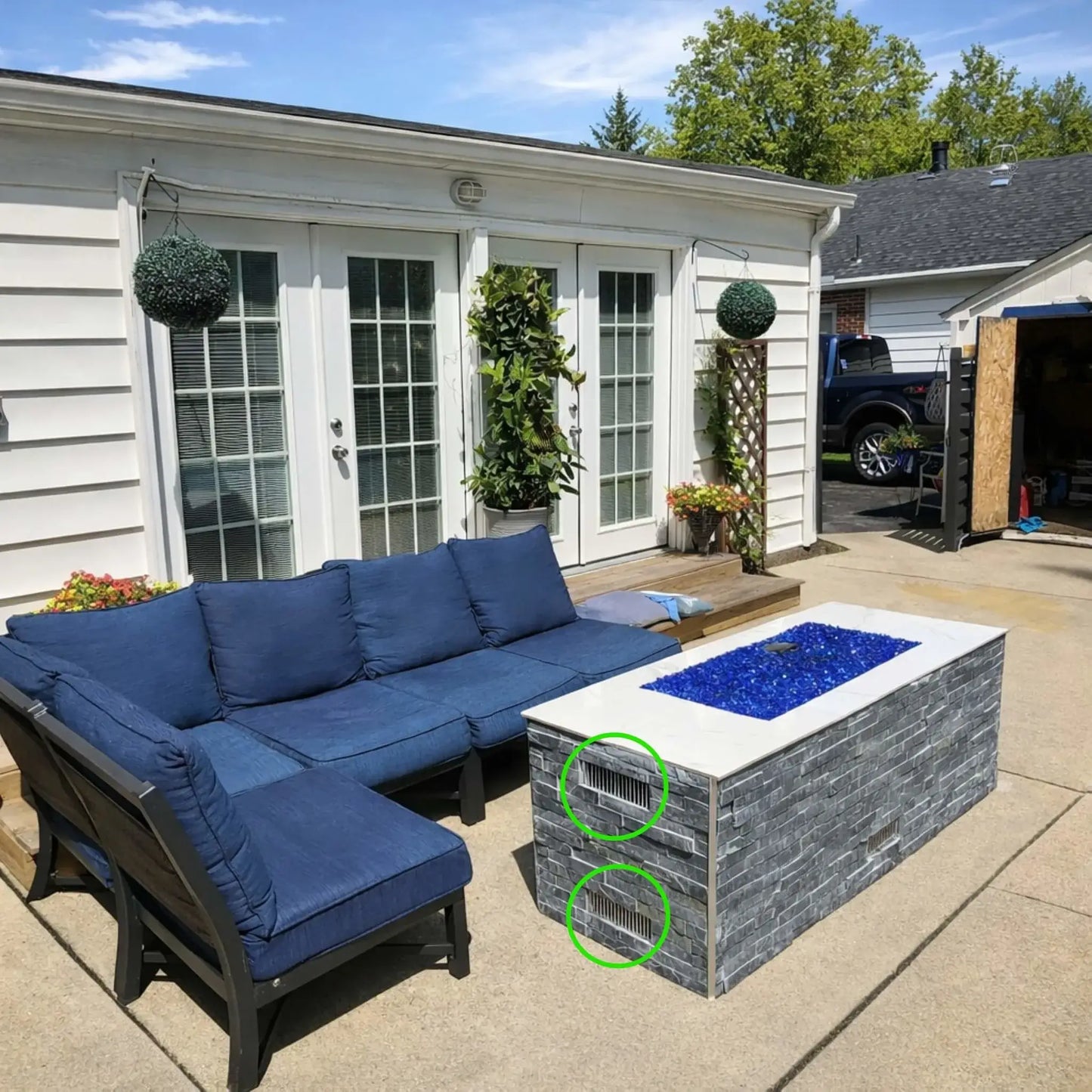 Backyard patio with sectional outdoor seating and a rectangular gas fire pit table filled with bright blue fire glass. Two stainless steel ventilation grilles are installed on the fire pit enclosure walls to provide proper airflow and gas ventilation for safe operation of the outdoor fire feature.