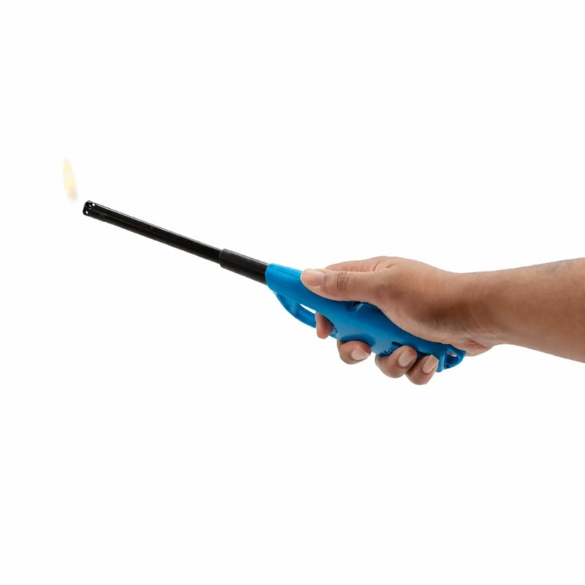 Hand holding a blue long-reach butane lighter igniting a small flame at the tip, designed for safely lighting outdoor fire pits, fireplaces, and gas burners. The extended black nozzle allows precise ignition from a distance, making it ideal for match-lit fire pit systems and manual lighting applications.