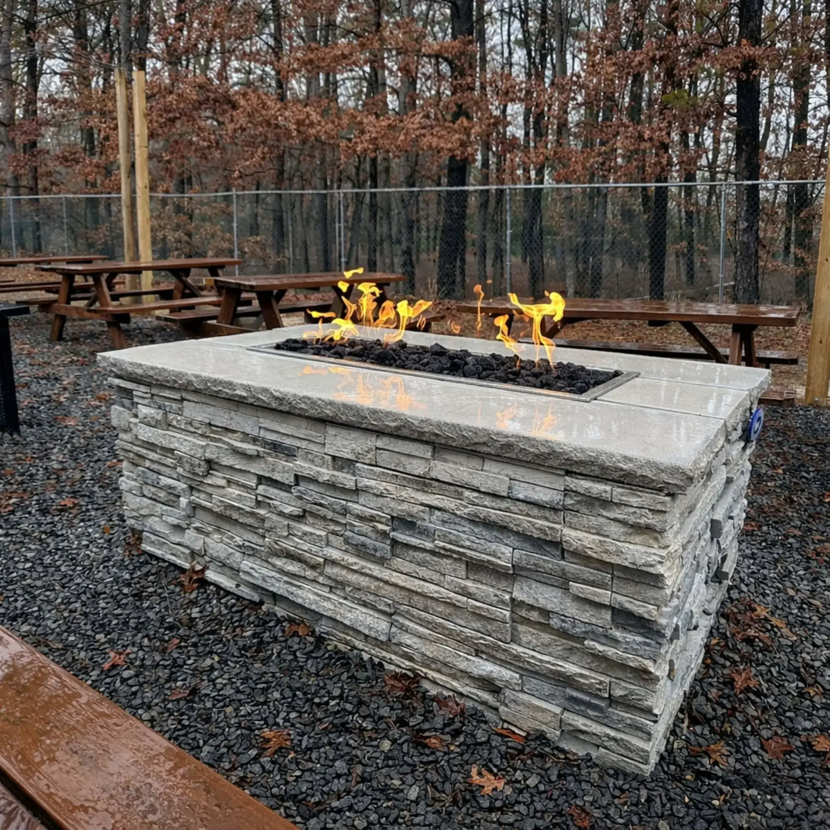 Hearth Products Controls HPC Fire rectangle drop in pan H burner torpedo electronic ignition Hi Lo low linear fire table built into a stone structure in a wooded outdoor picnic area, featuring black lava rock and natural flame pattern, controlled with a remote or app offering adjustable high and low flame settings.