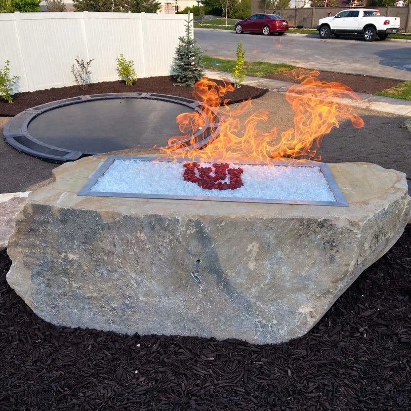 Hearth Products Controls HPC Fire rectangle drop in pan H burner torpedo electronic ignition Hi Lo low custom rock fire pit installation with white and red fire glass, set in a landscaped backyard with trampoline, demonstrating bold flame movement and easy remote or app control with high and low options.