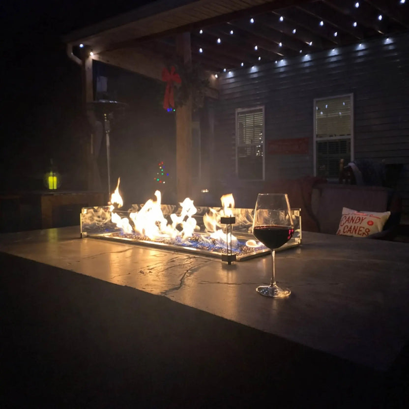 Hearth Products Controls HPC Fire rectangle drop in pan H burner torpedo electronic ignition Hi Lo low modern outdoor patio at night with linear fire feature, glass wind guard, and wine glass nearby, showing ambient flame lighting and simple control via remote or smartphone app with adjustable high and low flame levels.