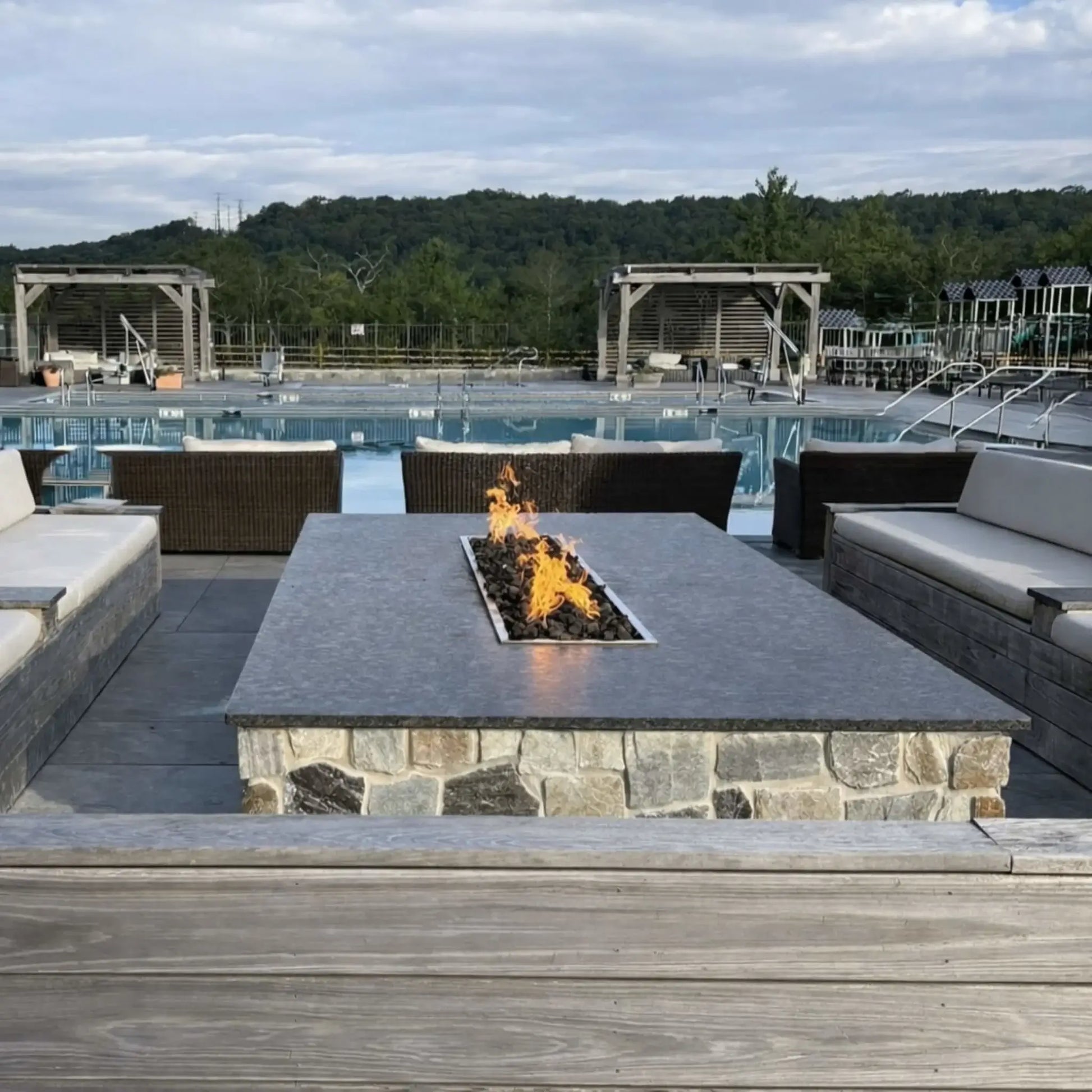 Hearth Products Controls HPC Fire rectangle drop in pan H burner torpedo electronic ignition Hi Lo low large outdoor fire table centered in a luxury poolside seating area with stone base and dark fire media, creating a warm focal point, with convenient control through remote or smartphone app offering high and low flame adjustments.