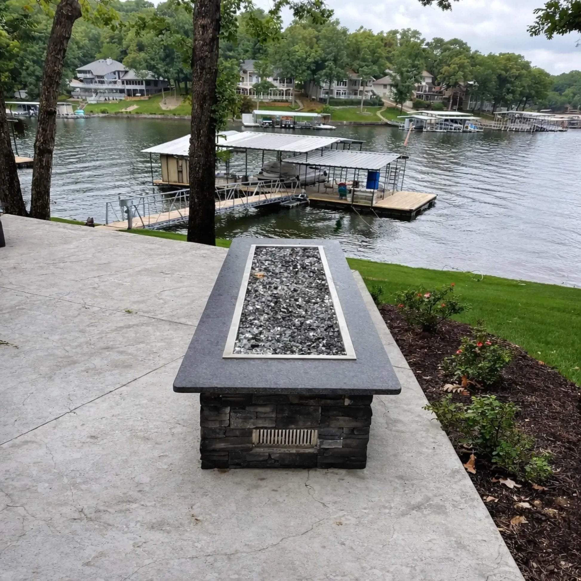 Hearth Products Controls HPC Fire rectangle drop in pan H burner torpedo electronic ignition Hi Lo low unlit fire table overlooking a lake dock and waterfront homes, showing stainless steel pan and media bed ready for use with remote or app control supporting high and low flame settings.
