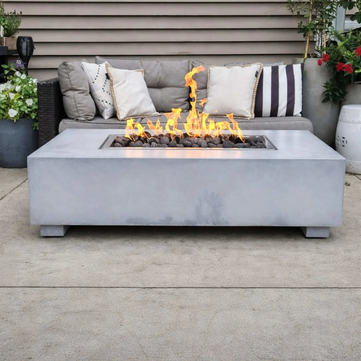 Stylish backyard patio showcasing Hearth Products Controls HPC Fire Rectangle Drop-In Pan H Burner Torpedo FPPK Push Button Flame Sensing FLEX fire table with smooth concrete finish, centered in front of a cushioned outdoor sofa with decorative pillows and potted plants.