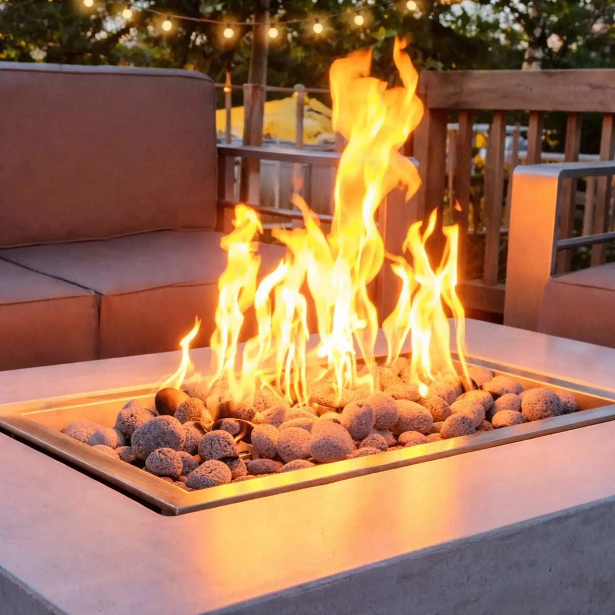 Close-up of a rectangular fire table featuring the Hearth Products Controls HPC Fire Rectangle Drop-In Pan H Burner Torpedo FPPK Push Button Flame Sensing FLEX system, with bright flames rising over smooth lava rocks, surrounded by cushioned patio seating and warm string lights in a cozy backyard setting.