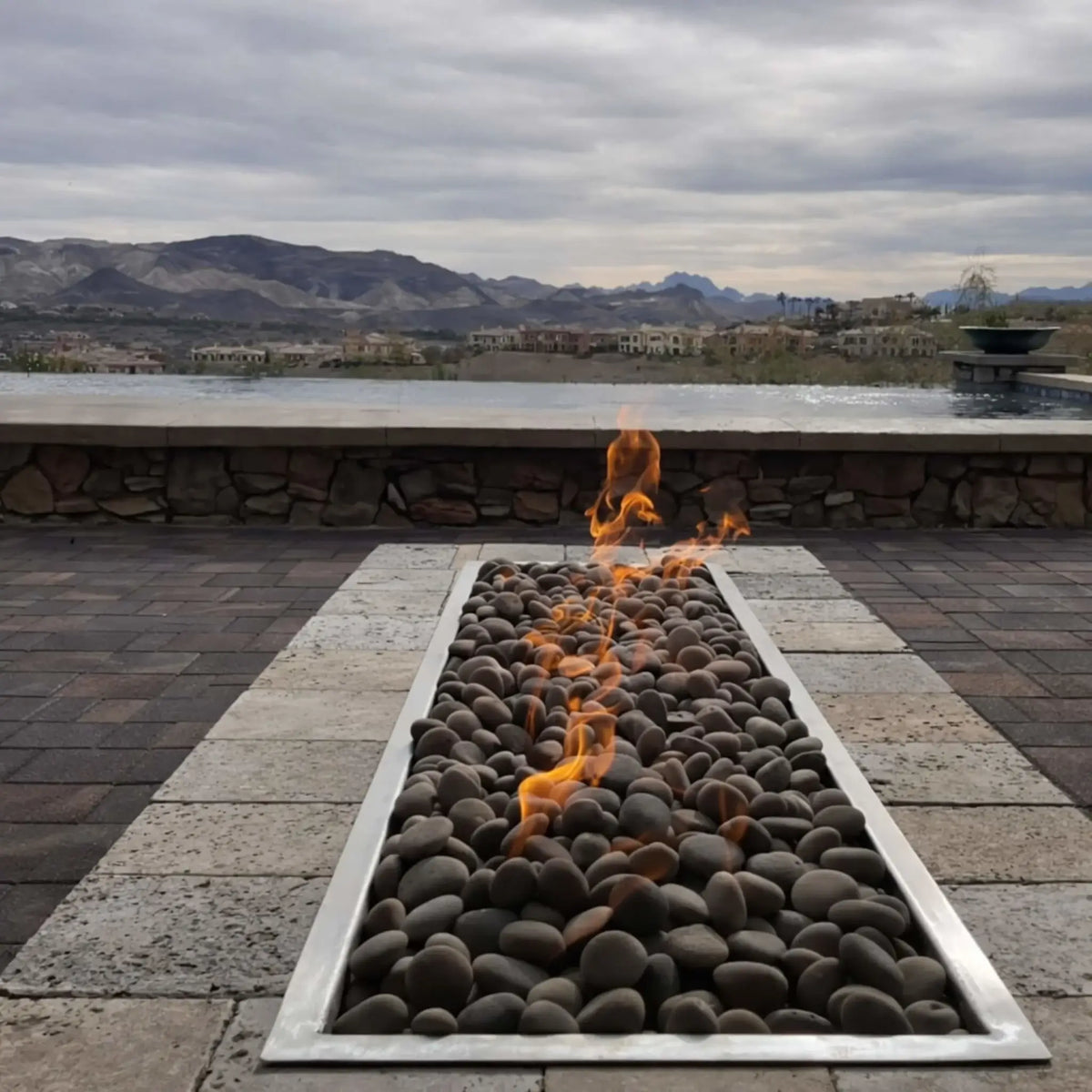 Modern commercial outdoor patio space featuring a large rectangular fire table equipped with Hearth Products Controls HPC Fire Rectangle Drop-In Pan H Burner Torpedo FPPK Push Button Flame Sensing FLEX system, surrounded by black metal seating and dining tables under a covered structure.