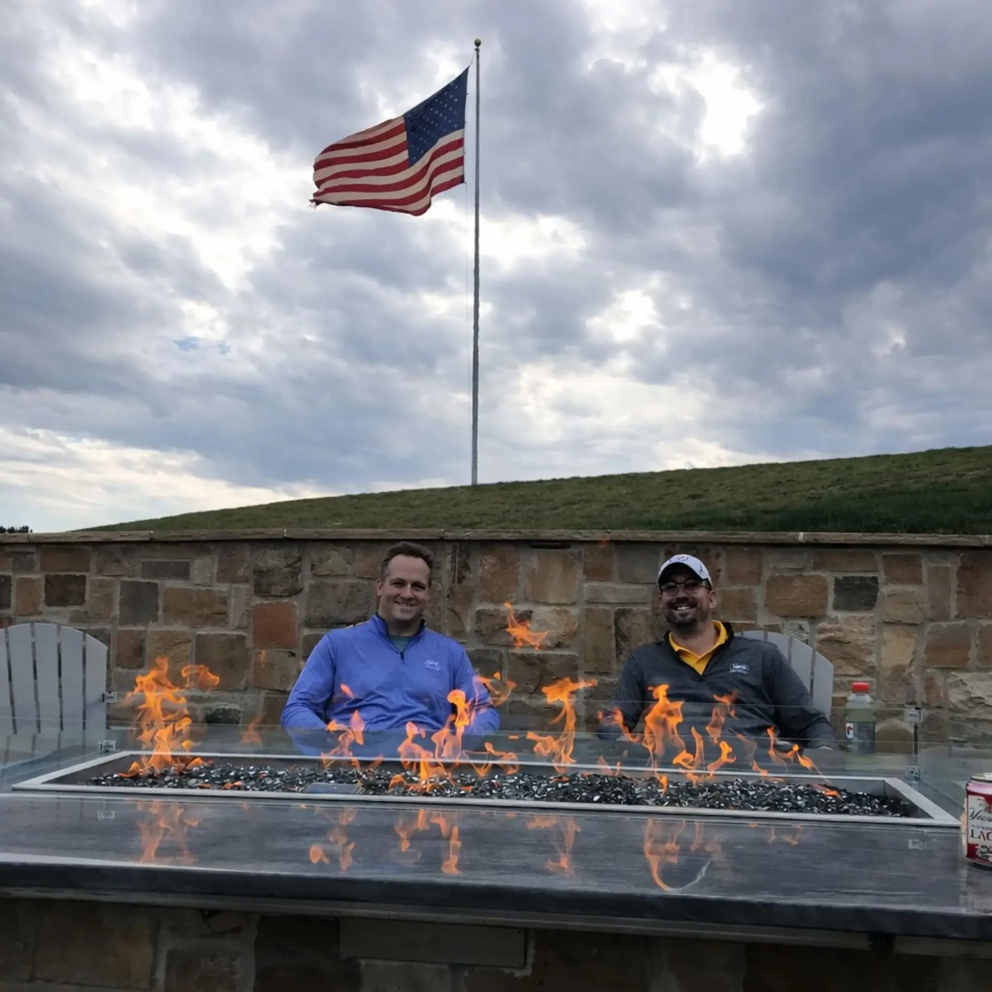 Two men relaxing behind a modern fire table featuring Hearth Products Controls HPC Fire Rectangle Drop-In Pan H Burner Torpedo FPPK Push Button Flame Sensing FLEX system, with flames dancing over fire glass and an American flag waving in the background.