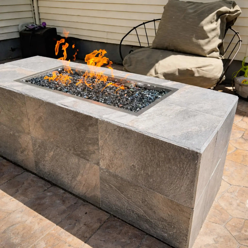 Contemporary rectangular stone fire table featuring Hearth Products Controls HPC Fire Rectangle Drop-In Pan H Burner Torpedo FPPK Push Button Flame Sensing FLEX burner, filled with dark fire glass and producing steady flames, positioned on a patio next to a cushioned lounge chair.