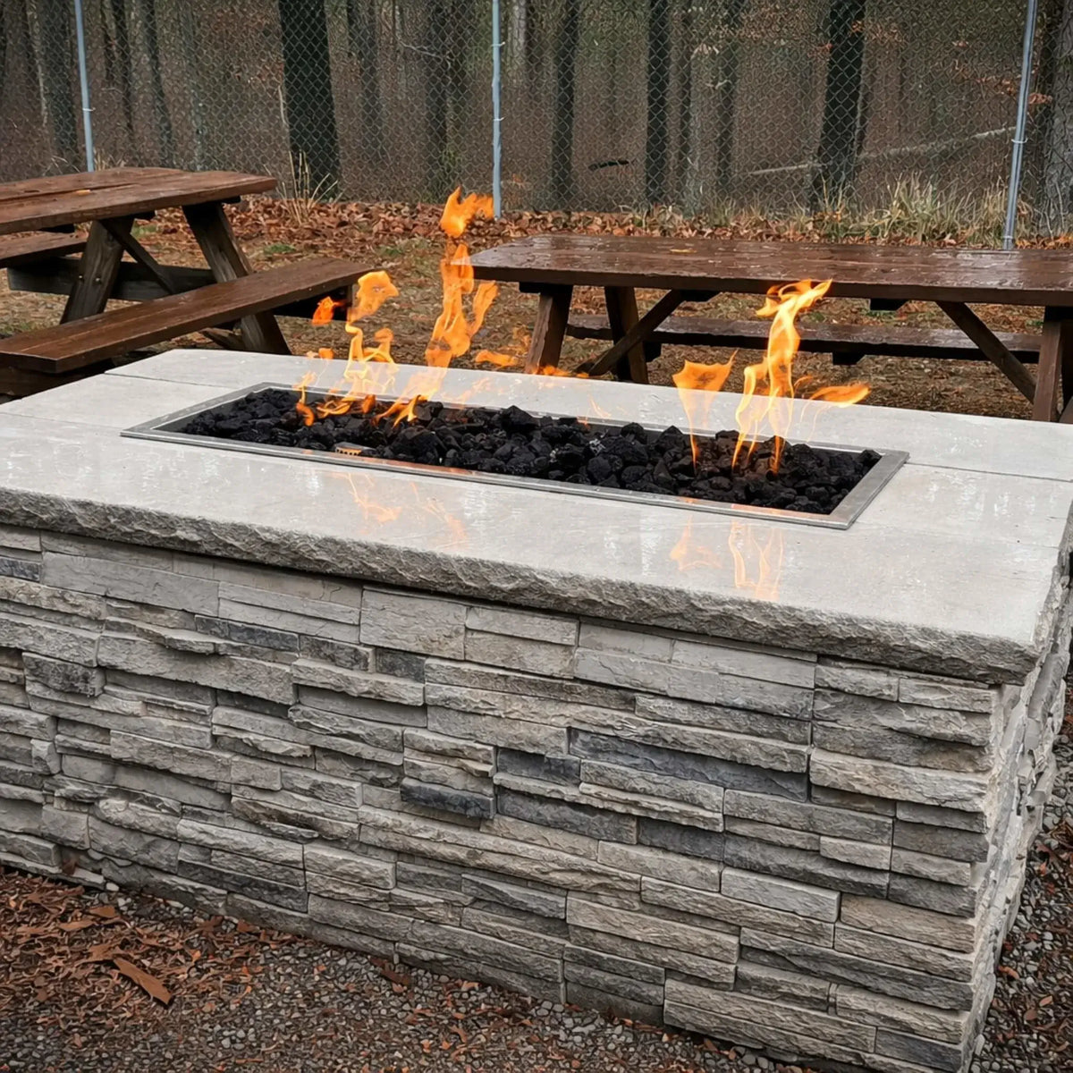 Linear stone fire table featuring Hearth Products Controls HPC Fire Rectangle Drop-In Pan H Burner Torpedo FPPK Push Button Flame Sensing FLEX burner filled with black lava rock, positioned near wooden picnic tables in an outdoor park-like setting with trees and fallen leaves.