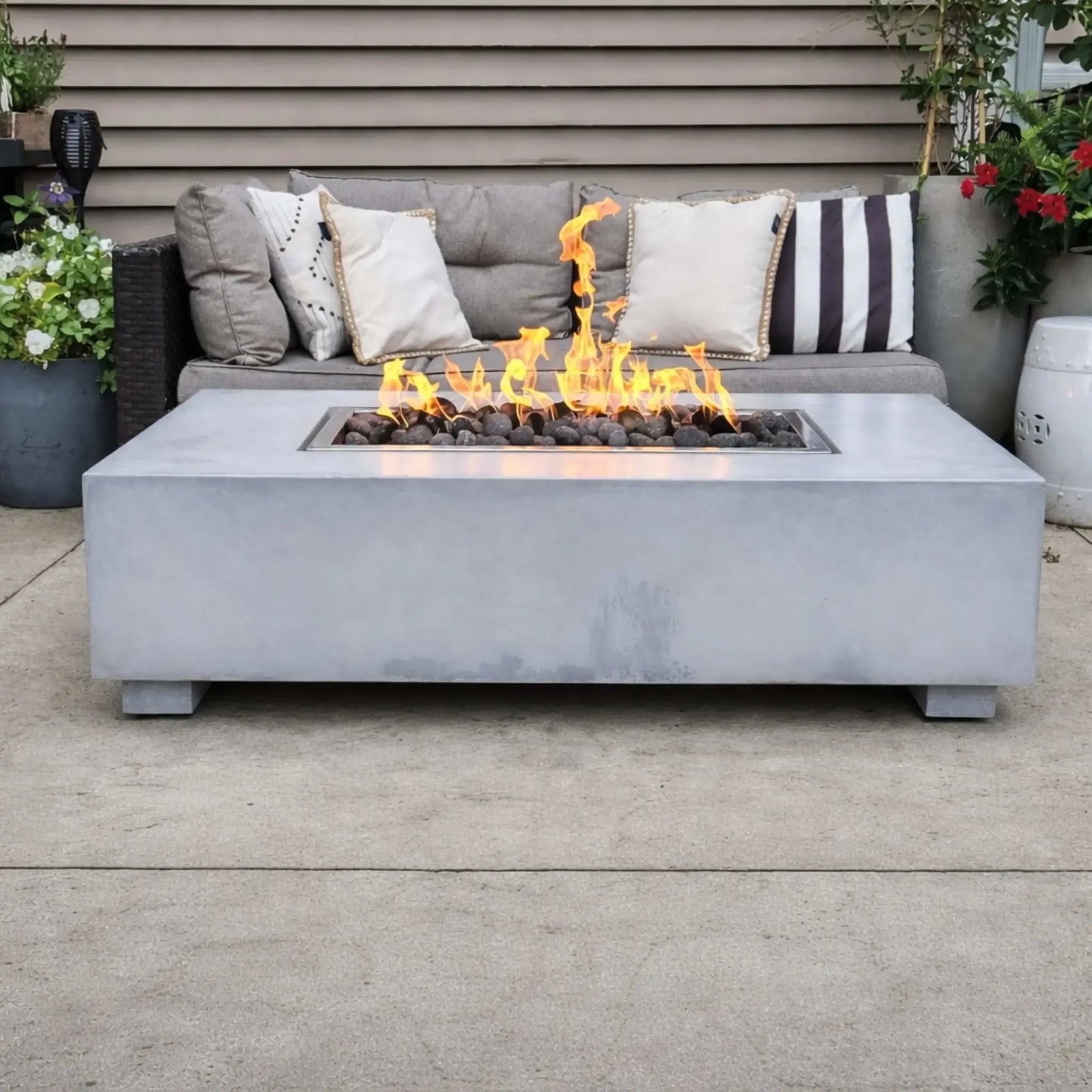 Concrete rectangular fire pit table with a stainless steel drop-in burner pan filled with lava rock, producing steady flames in front of a cushioned outdoor sofa. Decorative pillows and potted plants create a cozy backyard seating area with a warm, inviting fire feature centerpiece.
