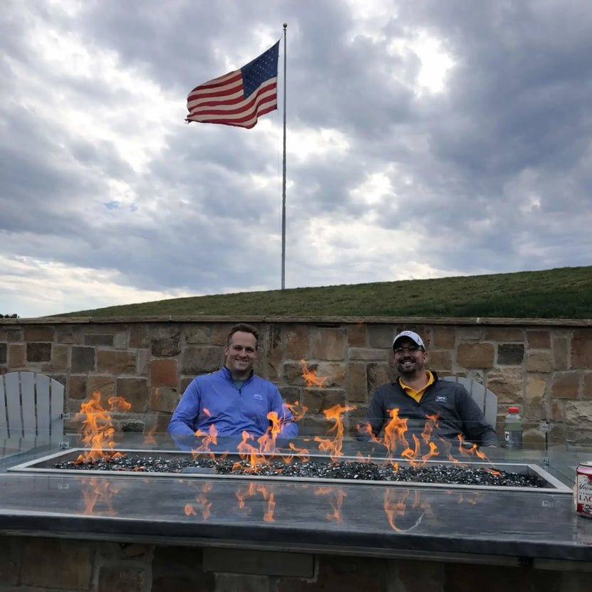 Two men seated behind a modern rectangular fire pit table with a glass wind guard, enjoying the flames in a backyard patio setting. An American flag waves in the background above a stone wall, highlighting a relaxed social atmosphere around a gas fire feature.