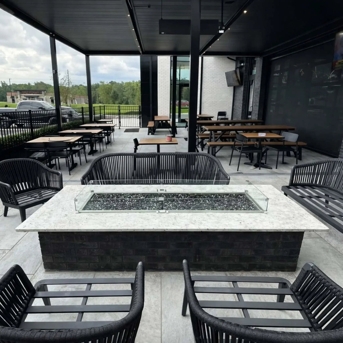 Covered commercial-style outdoor patio with multiple tables and chairs surrounding a central rectangular fire pit table. The burner is filled with dark fire media and enclosed by a glass wind guard, creating a stylish and functional gathering space for restaurants or lounges.