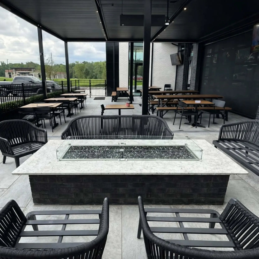 Covered commercial-style outdoor patio with multiple tables and chairs surrounding a central rectangular fire pit table. The burner is filled with dark fire media and enclosed by a glass wind guard, creating a stylish and functional gathering space for restaurants or lounges.