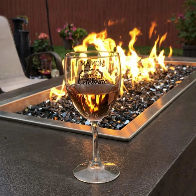 Close-up of a lit rectangular fire pit burner filled with black fire glass, with a wine glass in the foreground reflecting the flames. The scene showcases a cozy backyard setting with patio furniture, emphasizing the ambiance and warmth created by a gas fire pit insert.