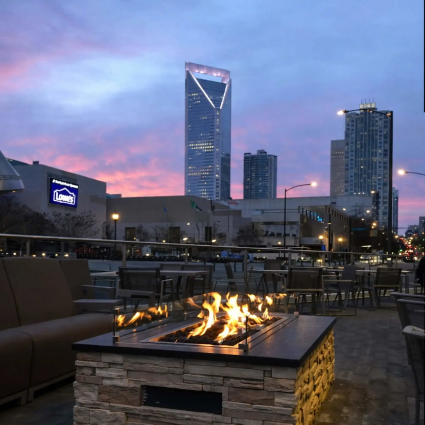 Modern rectangular fire pit table with a gray stone finish and drop-in burner filled with black fire glass, producing lively flames. Positioned on a backyard patio near a cushioned lounge chair, this setup showcases a compact and contemporary outdoor fire feature.
