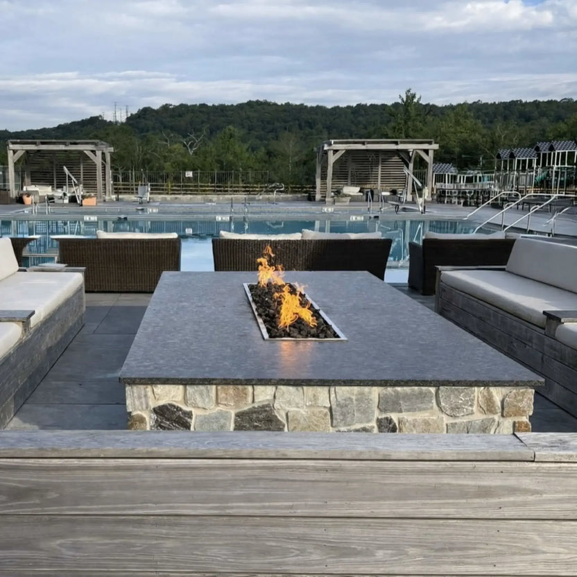Modern rectangular gas fire pit table with a stainless steel drop-in pan burner filled with dark lava rock, producing bright yellow flames at the center. The stone-clad base and smooth gray tabletop are surrounded by cushioned outdoor seating. Set beside a resort-style swimming pool with cabanas, lounge areas, and a wooded hillside backdrop under a cloudy sky, creating a relaxed, upscale outdoor living atmosphere.