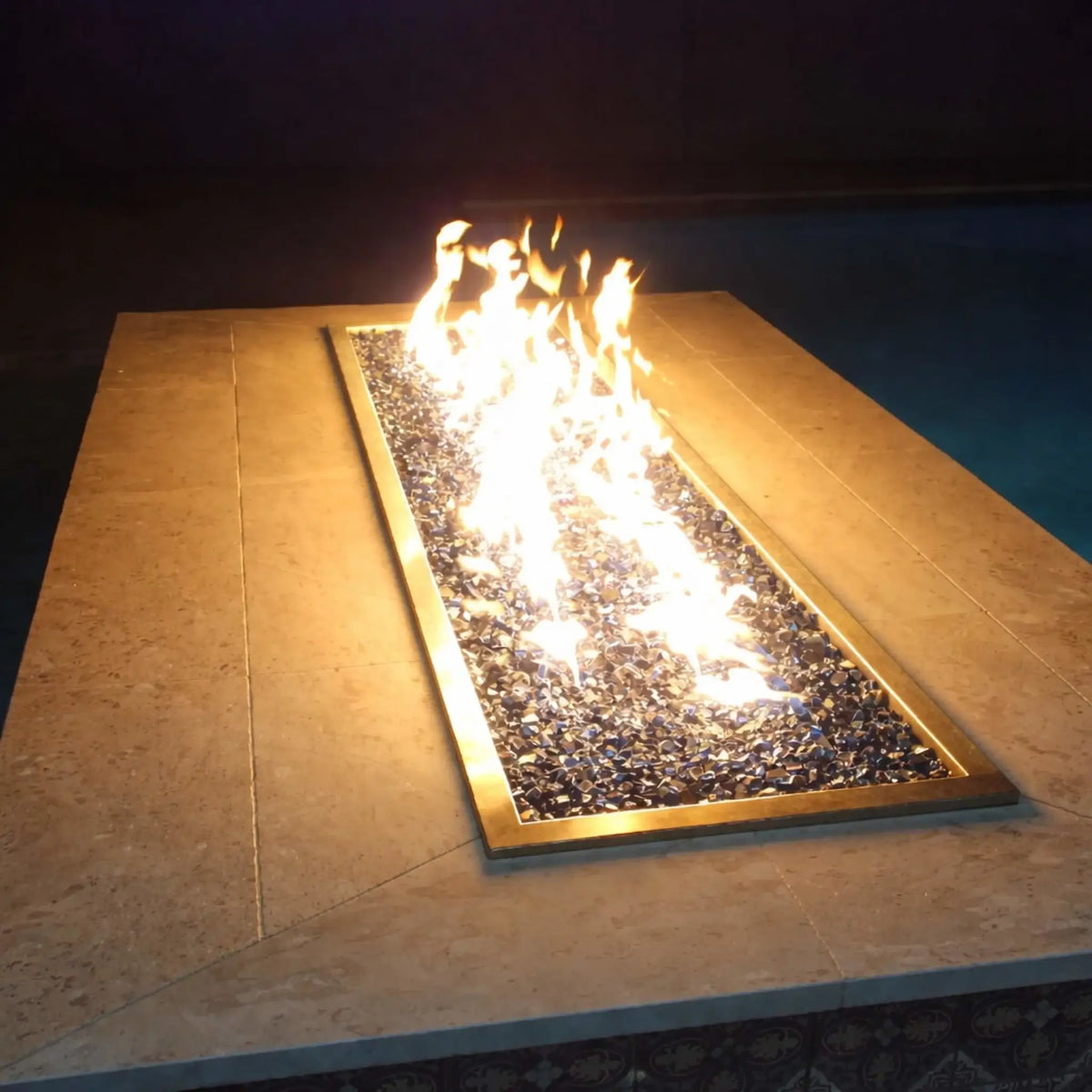 Nighttime close-up of Hearth Products Controls HPC Fire Rectangle Drop-In Pan H Burner Torpedo Match Lit system (MLFPK Flex) set in a stone fire table, showcasing bright flames over reflective fire glass, with a dark poolside background emphasizing the warm glow of the linear burner.