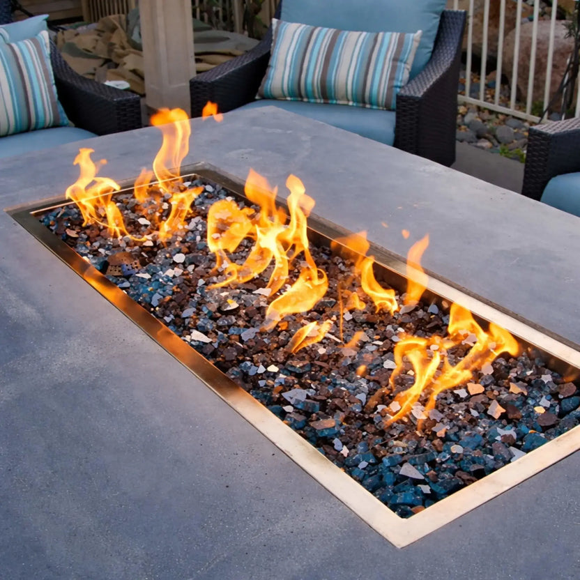 Rectangular HPC Fire Drop-In Pan H Burner Torpedo Match Lit fire feature (CSA MLFPK Flex) installed in a concrete patio table with mixed blue and black fire glass, surrounded by cushioned outdoor chairs in a modern backyard lounge setting.
