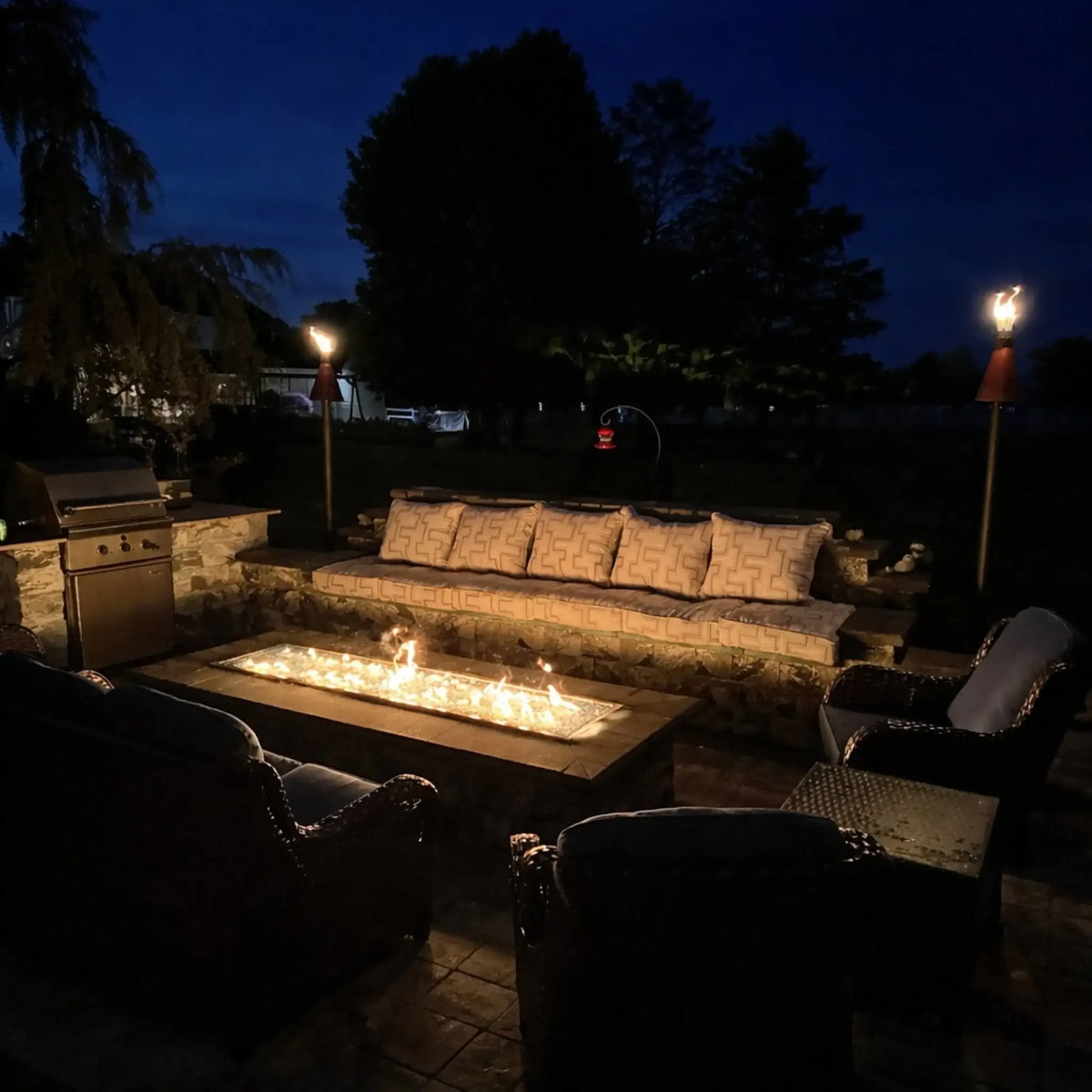 Nighttime outdoor living space featuring Hearth Products Controls HPC Fire Rectangle Drop-In Pan H Burner Torpedo Match Lit system (MLFPK Flex) in a long fire pit table, surrounded by wicker seating, patio lighting, and tiki torches creating a warm evening atmosphere.