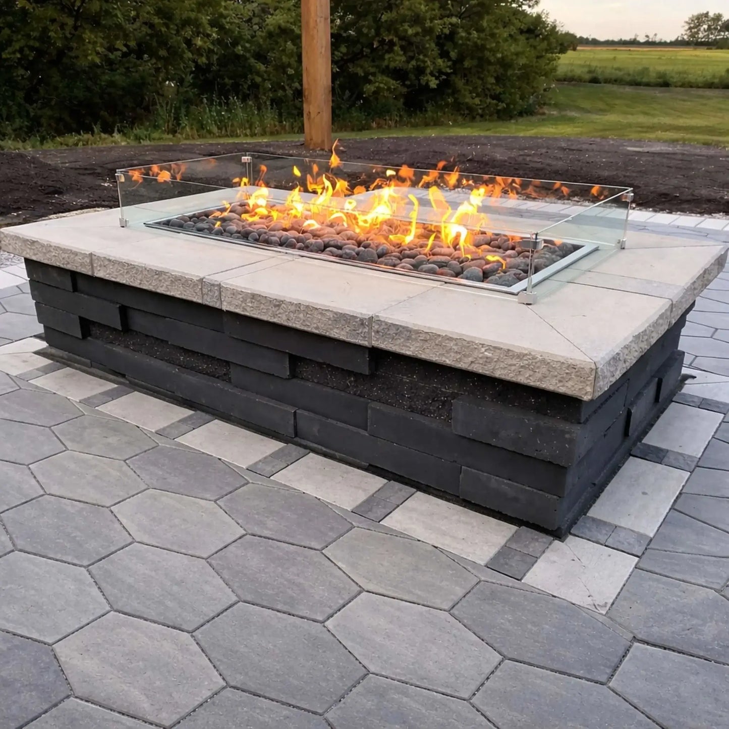 Contemporary outdoor fire pit installation using Hearth Products Controls HPC Fire Rectangle Drop-In Pan H Burner Torpedo Match Lit system (MLFPK Flex), featuring a glass wind guard, lava rock media, and a paved patio with landscaped yard and trees in the background.