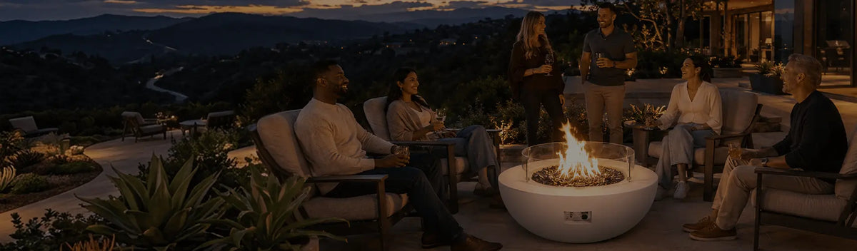 Family and friends gather around a white fire bowl overlooking a valley at sunset.