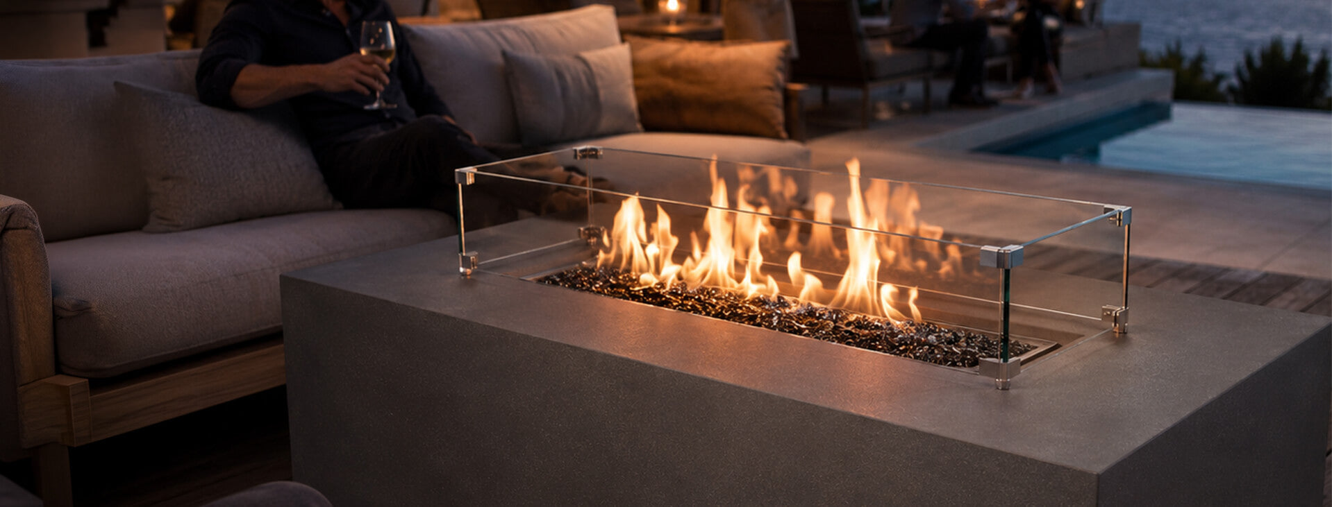 Built for performance lifestyle image of a tall rectangular gray concrete fire pit with lively flames rising through reflective black fire glass. A clear tempered glass wind guard surrounds the linear burner, while a guest relaxes with a glass of wine beside a swimming pool at dusk.