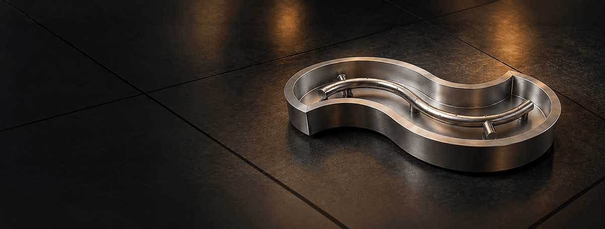 Custom burner banner for desktop featuring a sculpted stainless steel fire pit burner pan with a flowing, organic curved shape and matching tubular gas burner with evenly spaced ports. The polished metal assembly is displayed on dark stone tile under warm architectural lighting, with open negative space on the left for website text or promotional content.