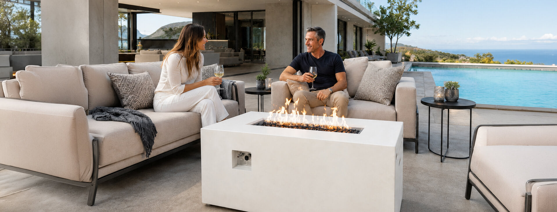 White rectangular fire pit lifestyle scene featuring a man and woman enjoying wine beside a modern linear fire table with bright flames and dark fire media. Neutral cushioned seating surrounds the tall rectangle fire pit on a luxury patio overlooking an infinity pool, rolling hills, and the blue ocean.