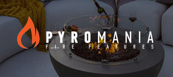 A round outdoor fire pit with a glass wind guard and visible flames, set in a patio environment with a yellow blanket and sofa in the background. A large 'PYROMANIA FIRE FEATURES' logo overlay is visible on the left.